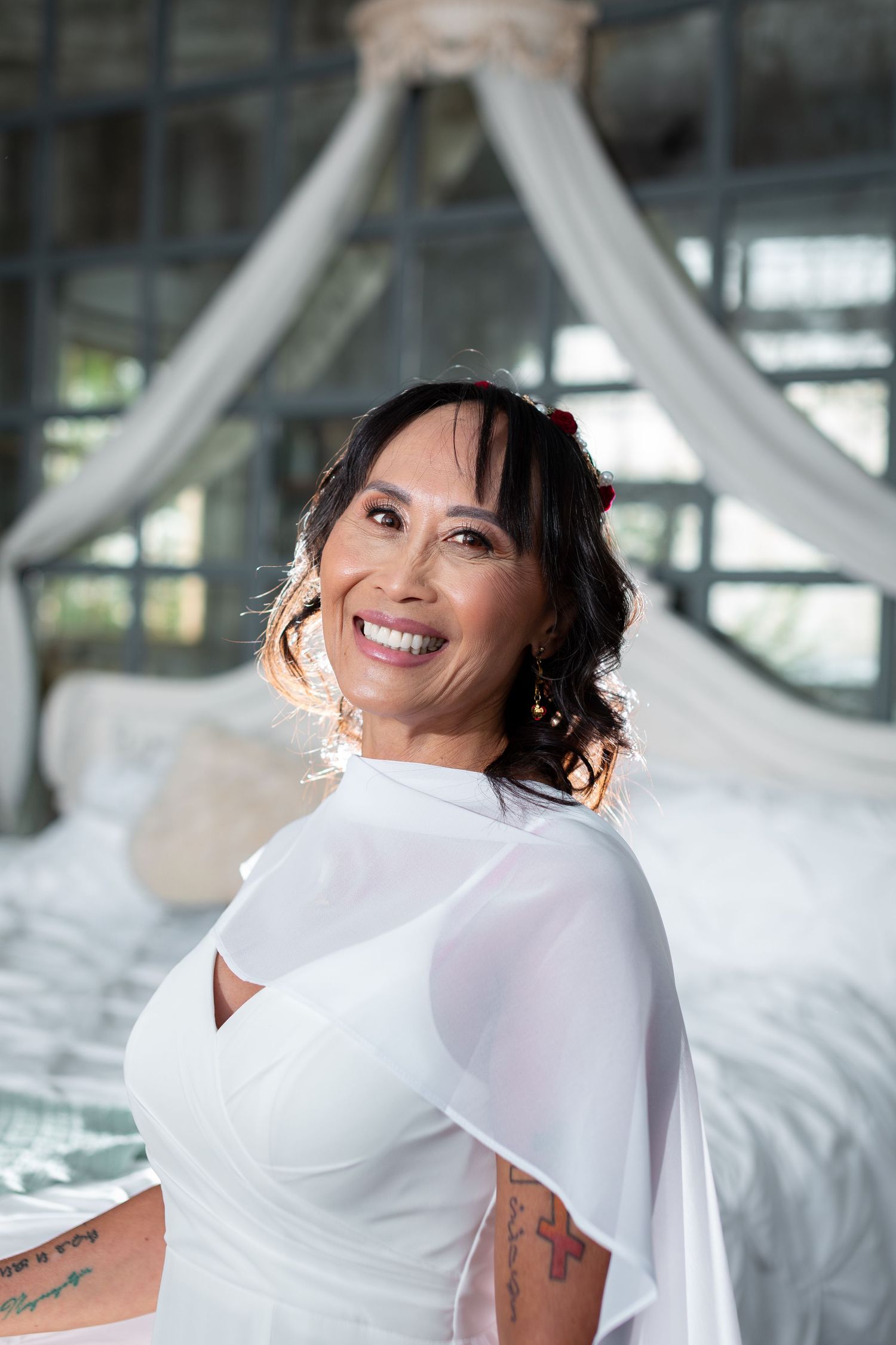 Smiling woman in white dress with sheer cape stands in elegant bedroom with canopy bed.