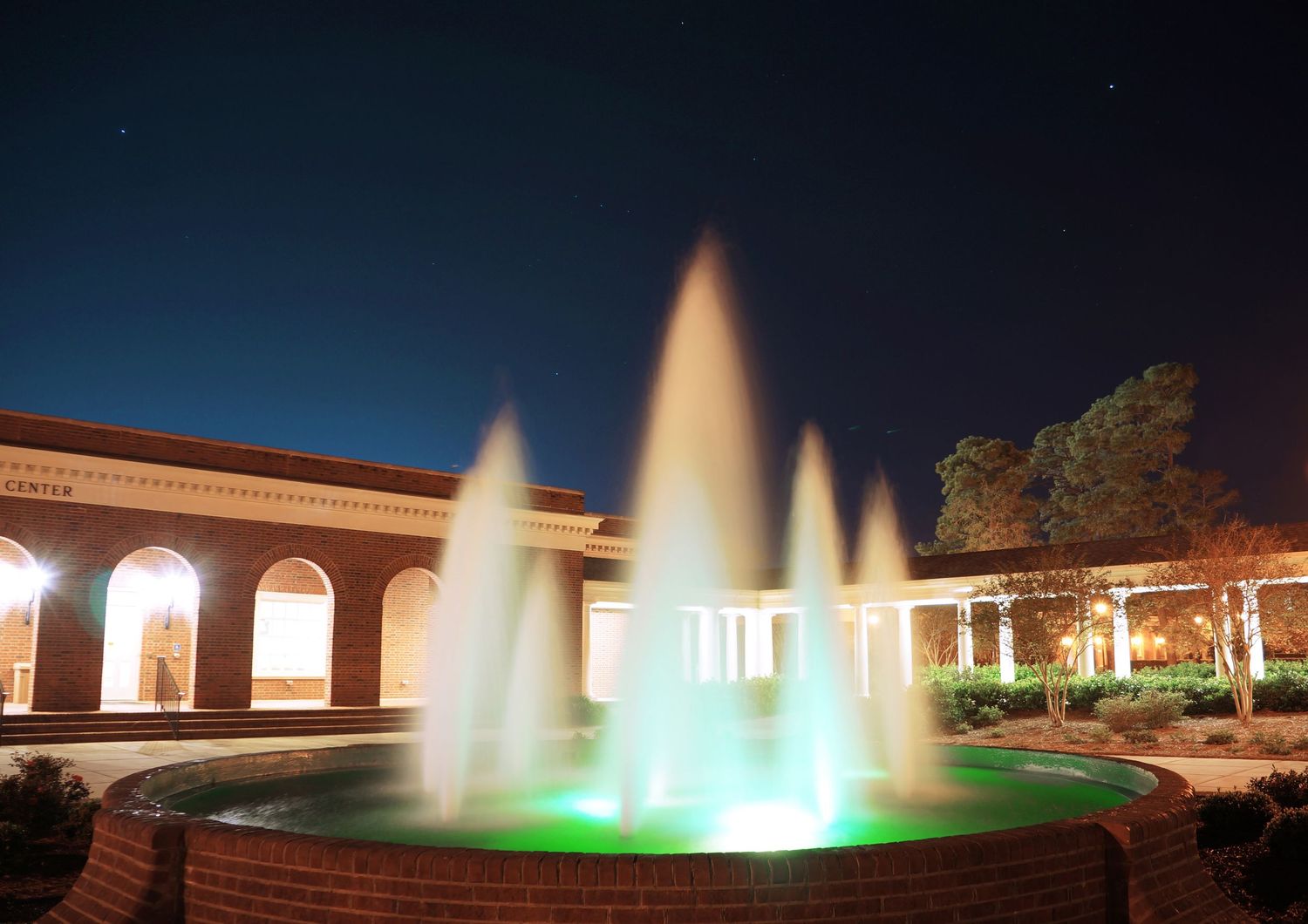 An illuminated outdoor fountain with green lights glows at night outside a brick building.