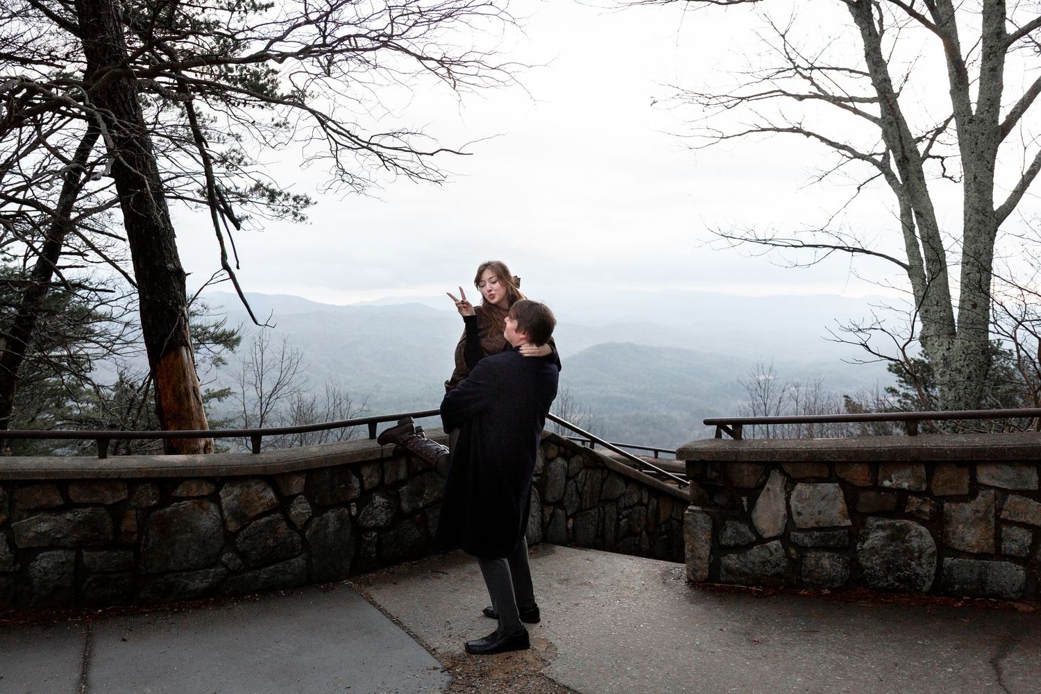 A silhouetted figure stands at a scenic mountain overlook with a stone wall and bare winter trees framing the view.