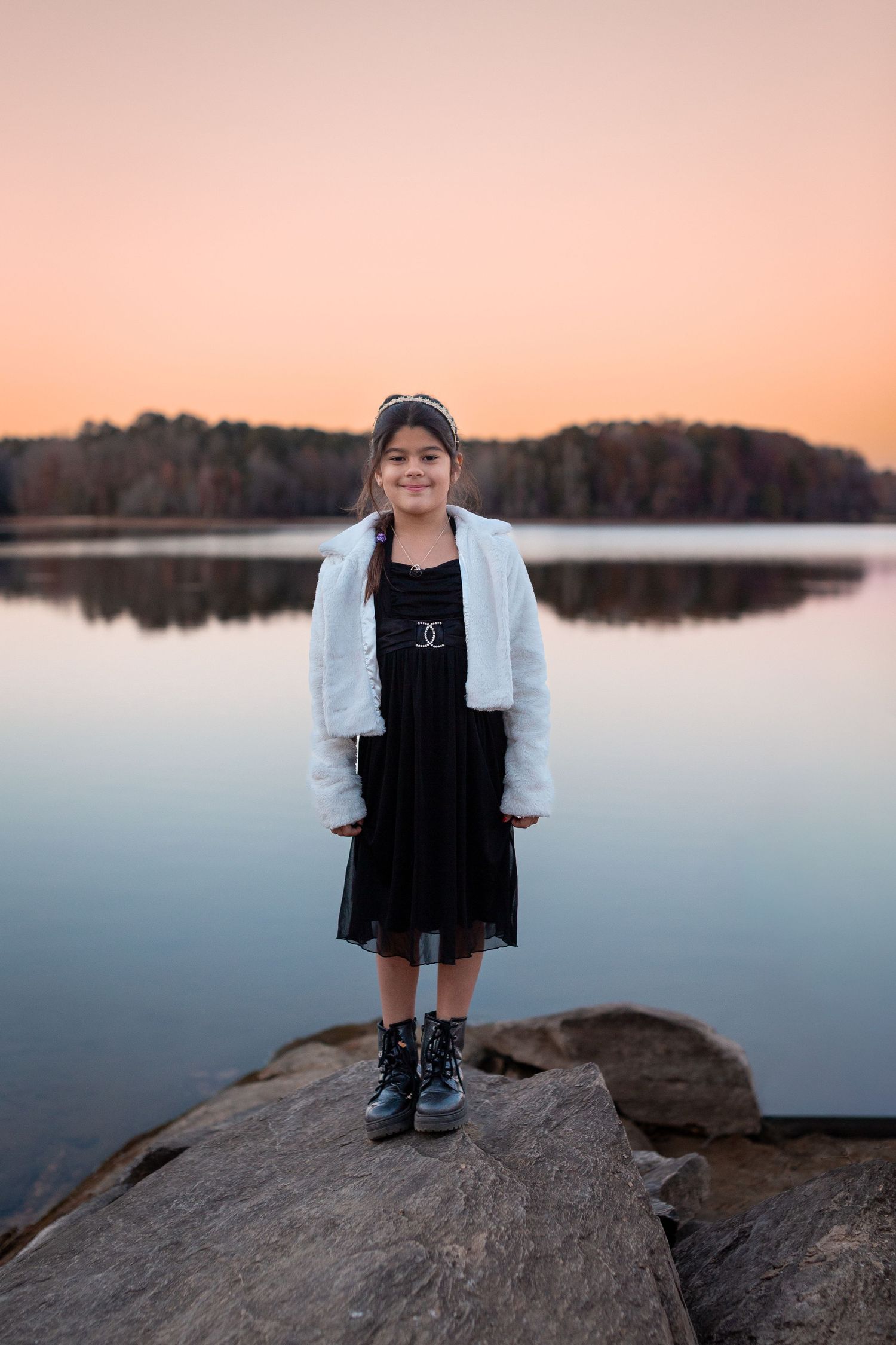 Child stands on rocky outcrop overlooking calm lake during sunset.