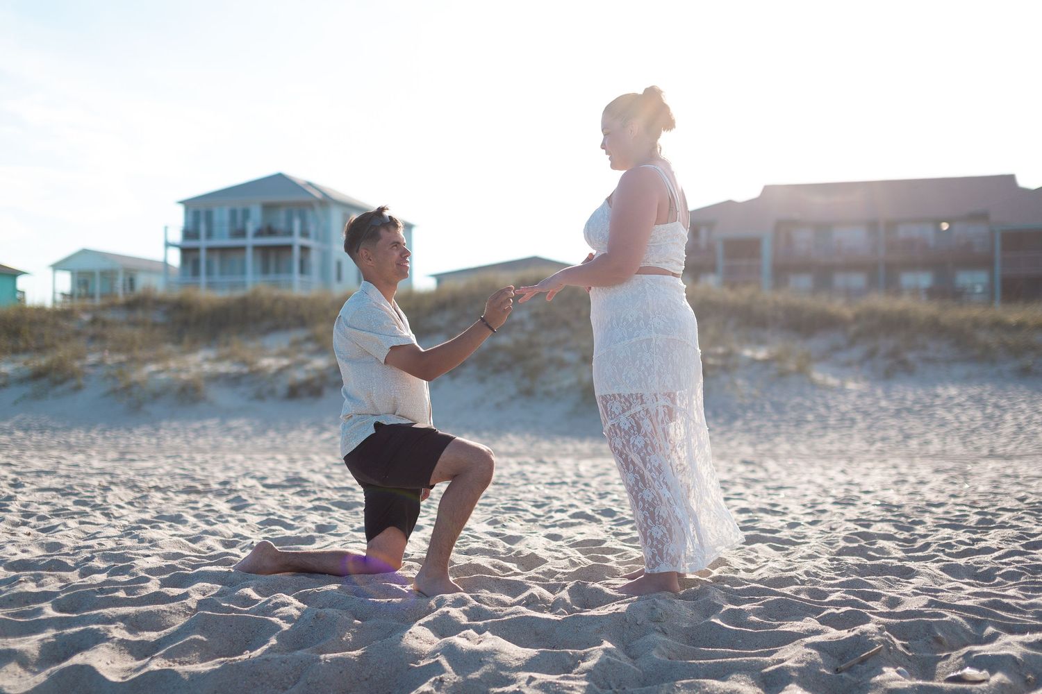 A romantic beach proposal at sunset with beach houses visible in the background.