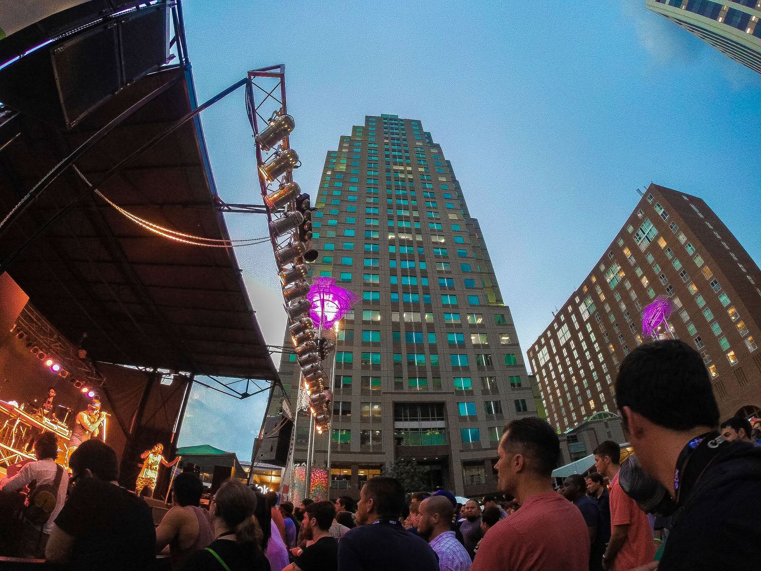 Tall urban buildings with glass facades are illuminated against a blue evening sky as crowds gather below.