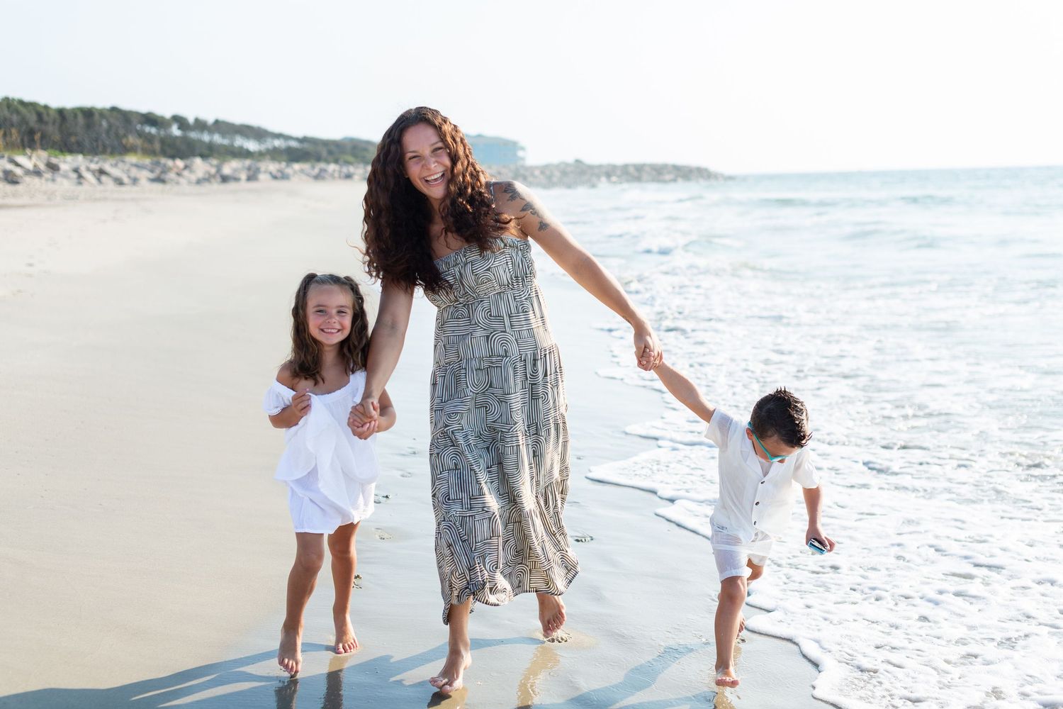 Candid family moments walking along beach shoreline in casual white outfits.
