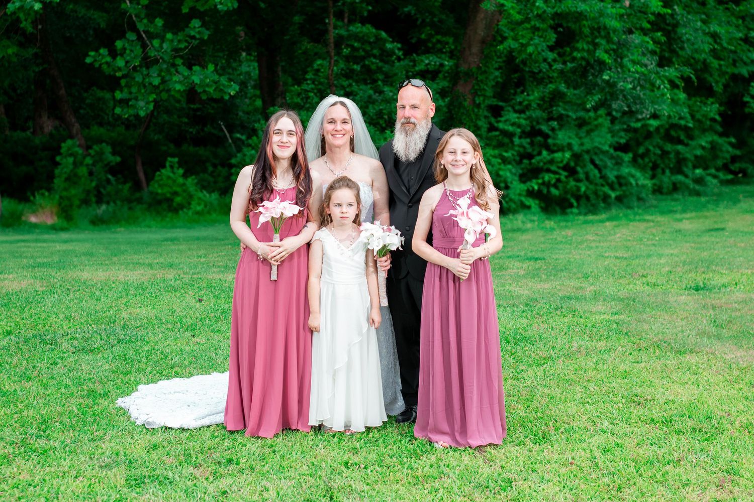 Wedding party in pink bridesmaid dresses poses together on lush green lawn during summer celebration.