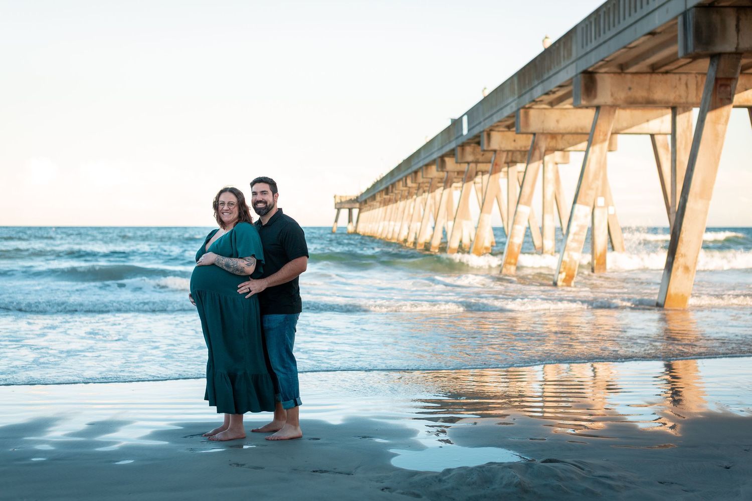 Expecting couple embracing barefoot on the beach near a wooden pier at sunset during a maternity photo session.