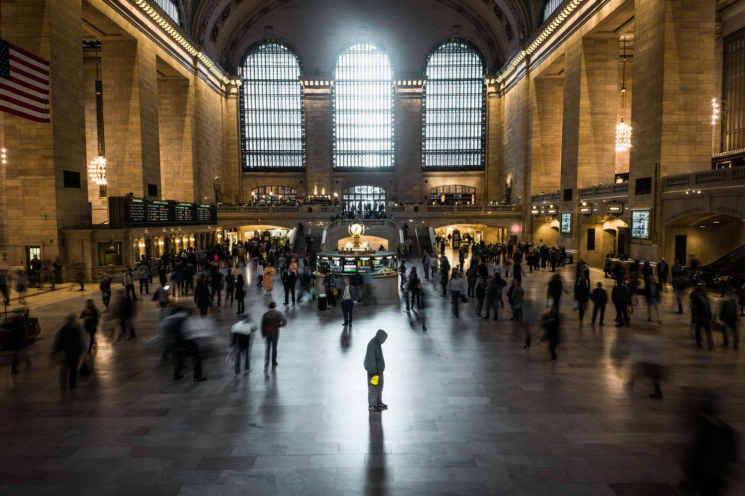 Busy Grand Central Terminal main concourse with crowds of commuters walking beneath grand arched windows and American flag.