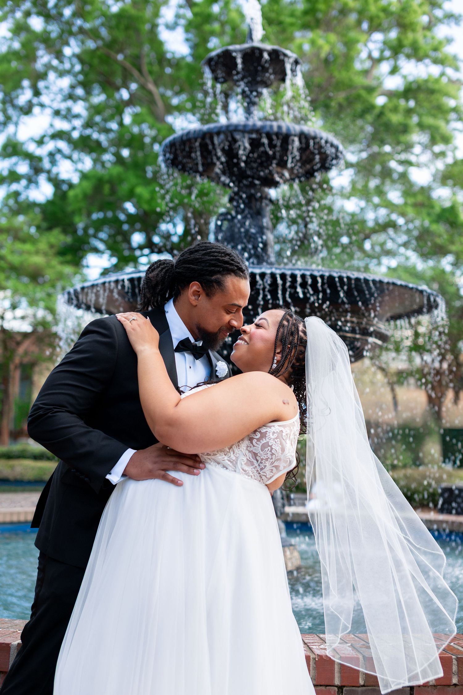 A Black bride and groom share a romantic moment in front of a tiered fountain, surrounded by lush green trees.