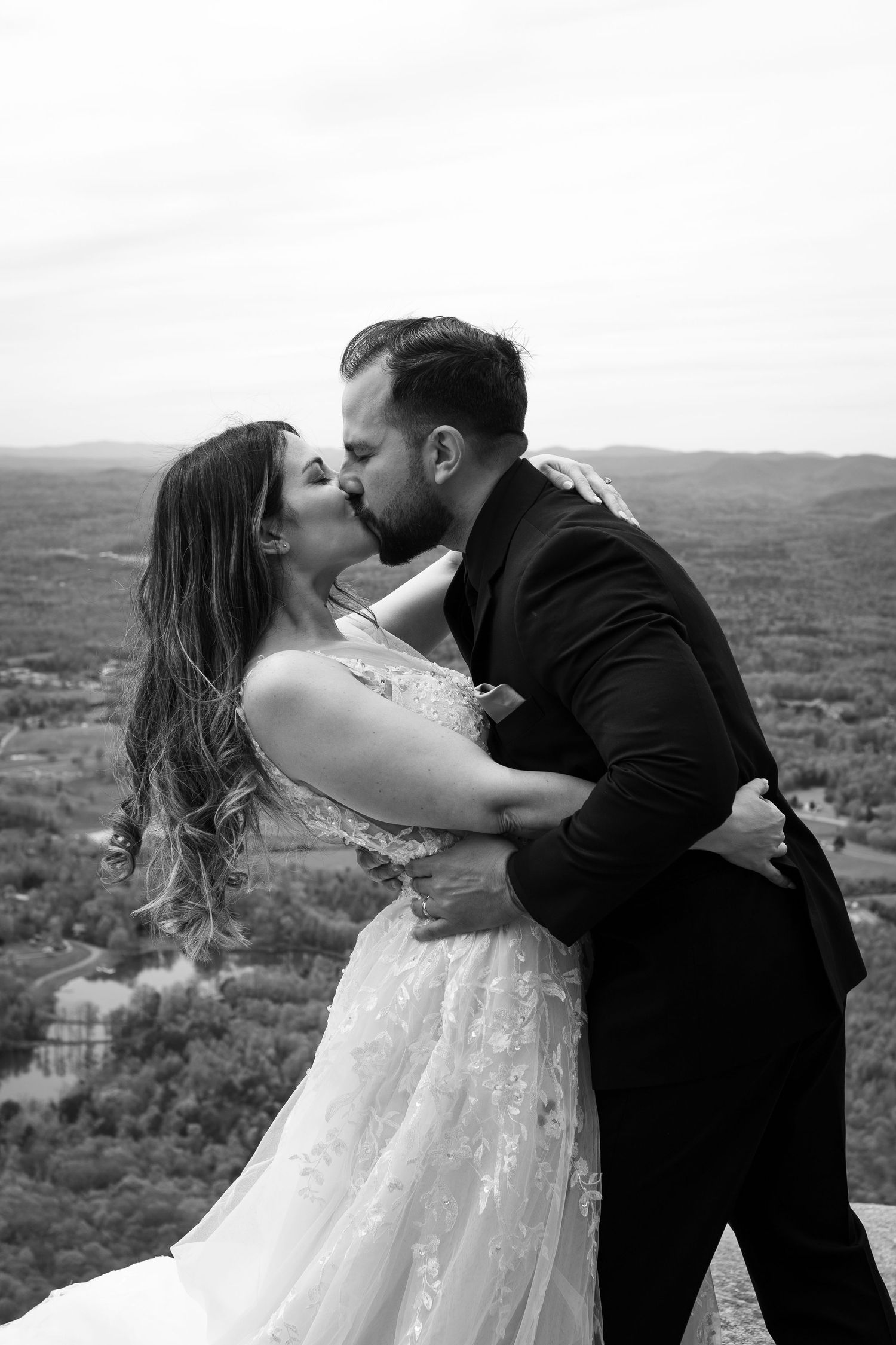 Black and white photo of a bride and groom sharing a romantic kiss with a scenic mountain valley backdrop.
