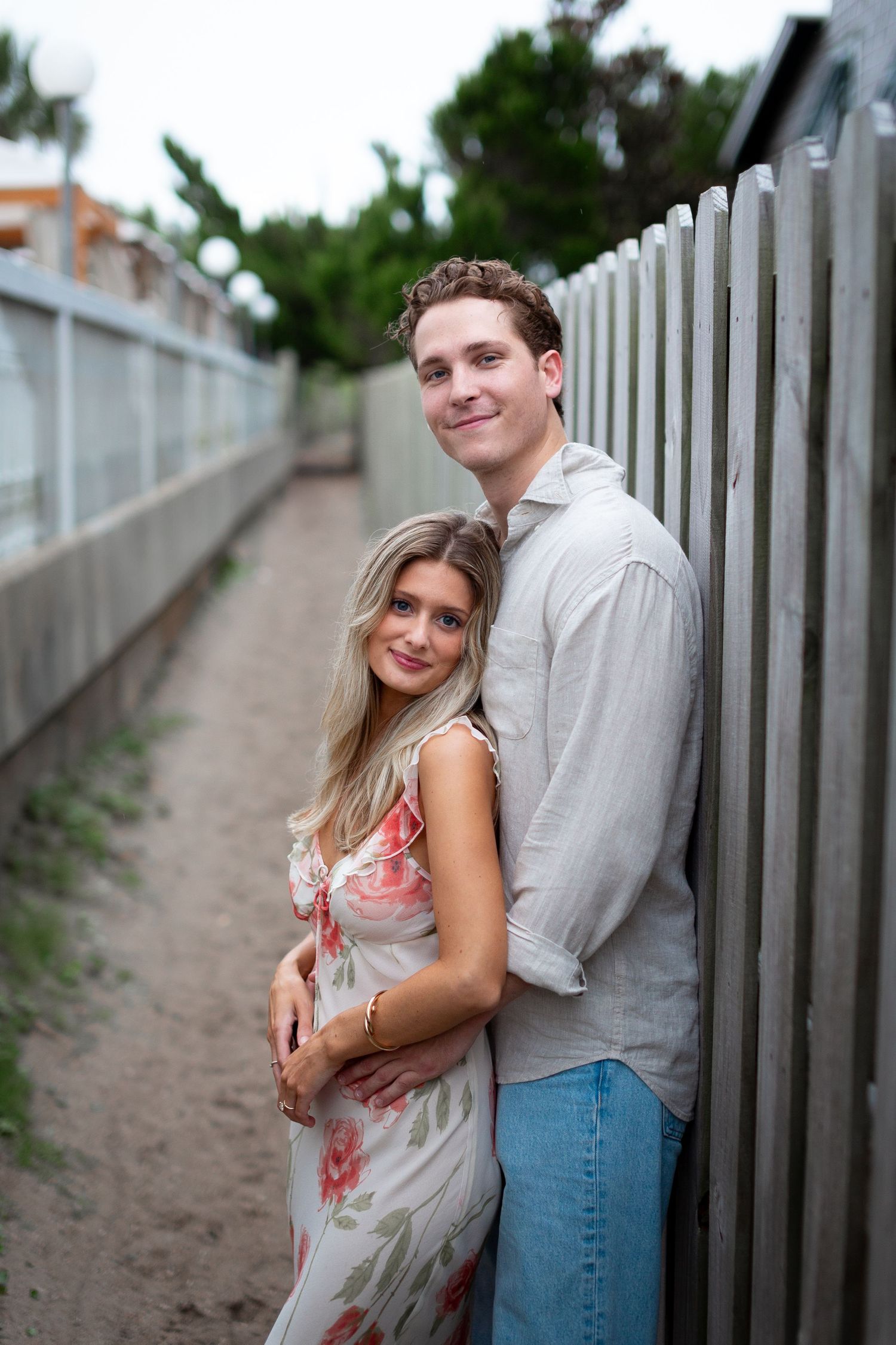 A couple posing against white fence posts along a beach walkway.