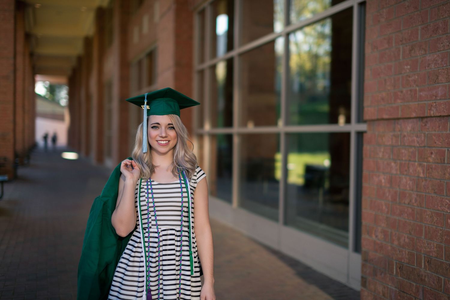 Graduate in green cap and gown poses on brick steps of a traditional collegiate building with white columns.