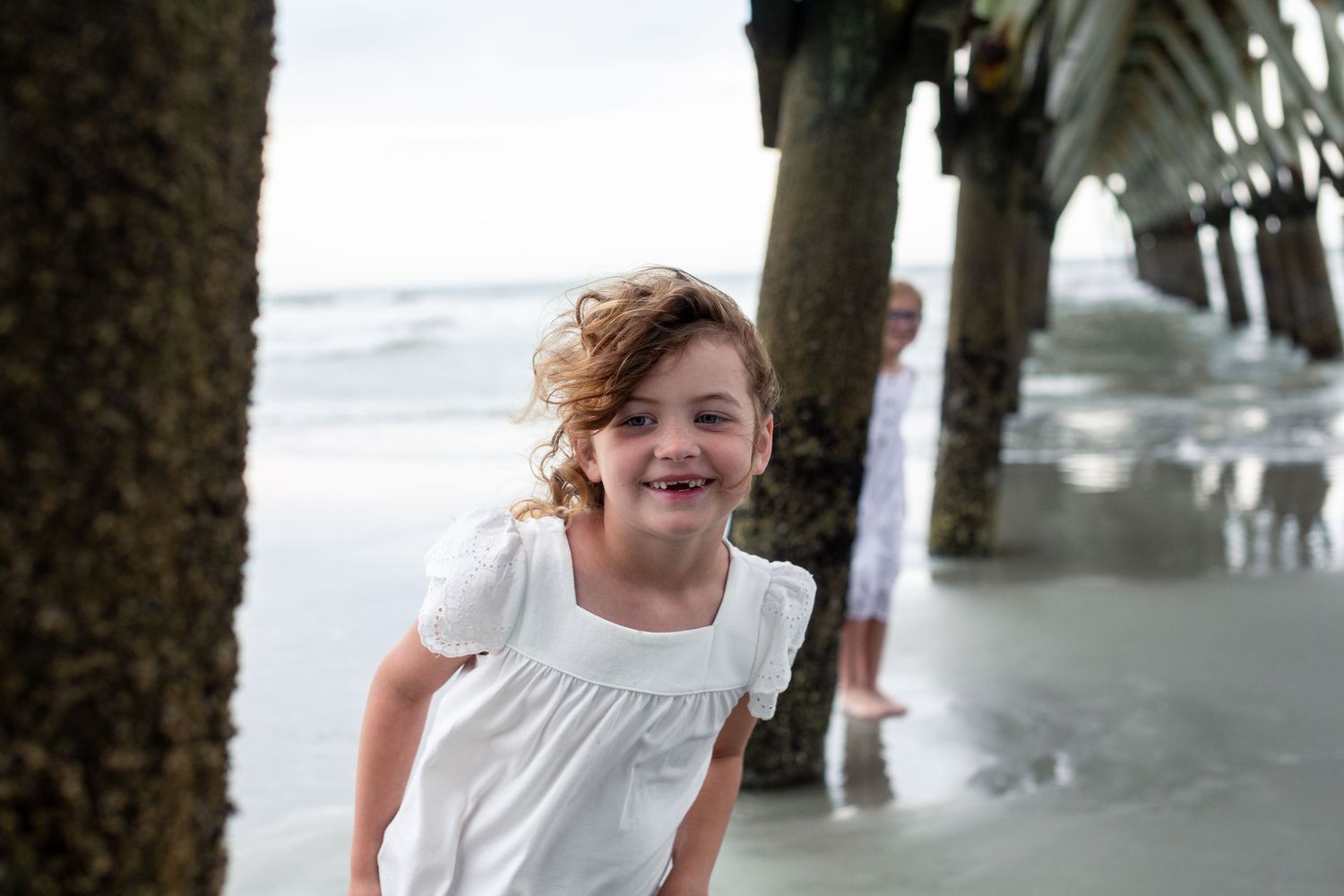 Child in a white top smiles joyfully near a pier at the beach.
