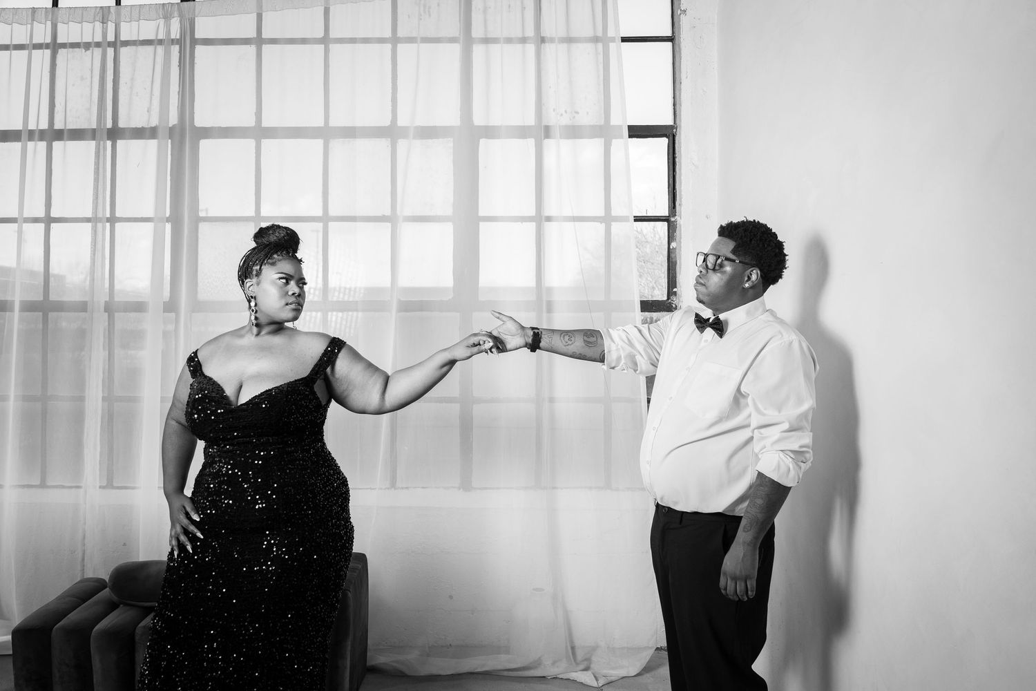 A black and white photo shows two people in elegant attire sharing a playful moment against a bright window backdrop.