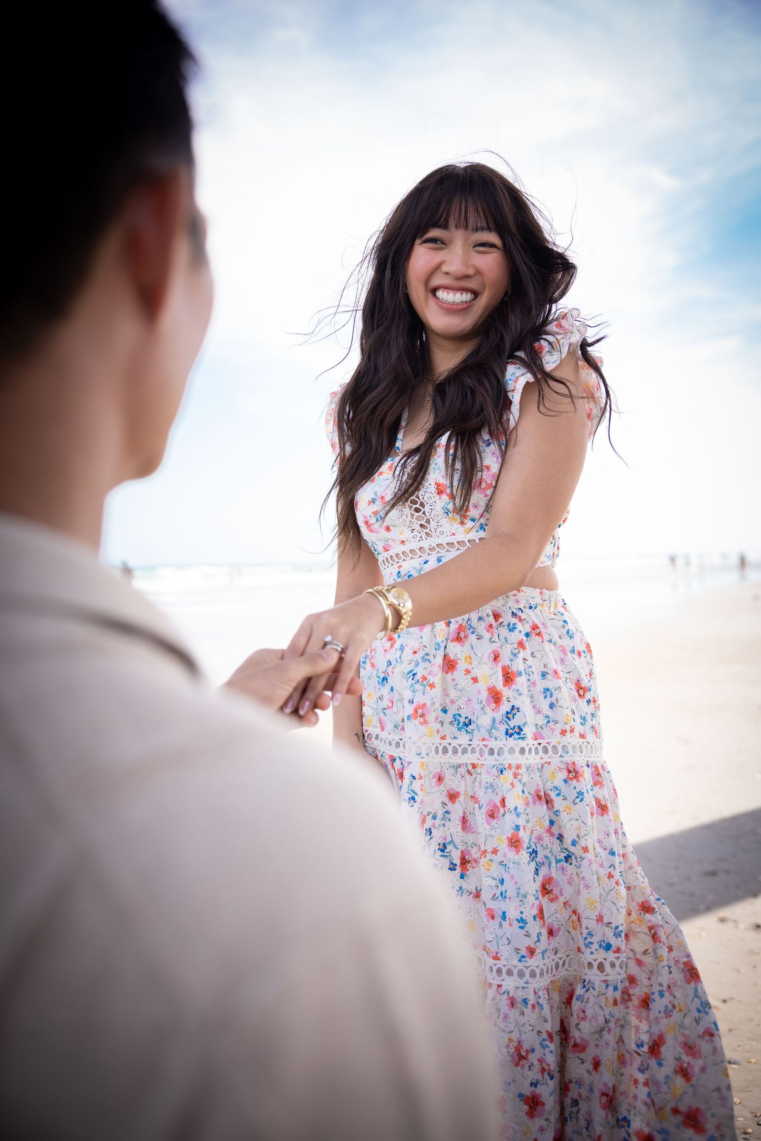Couple sharing joyful moment under bright sky with floral dress blowing in wind.