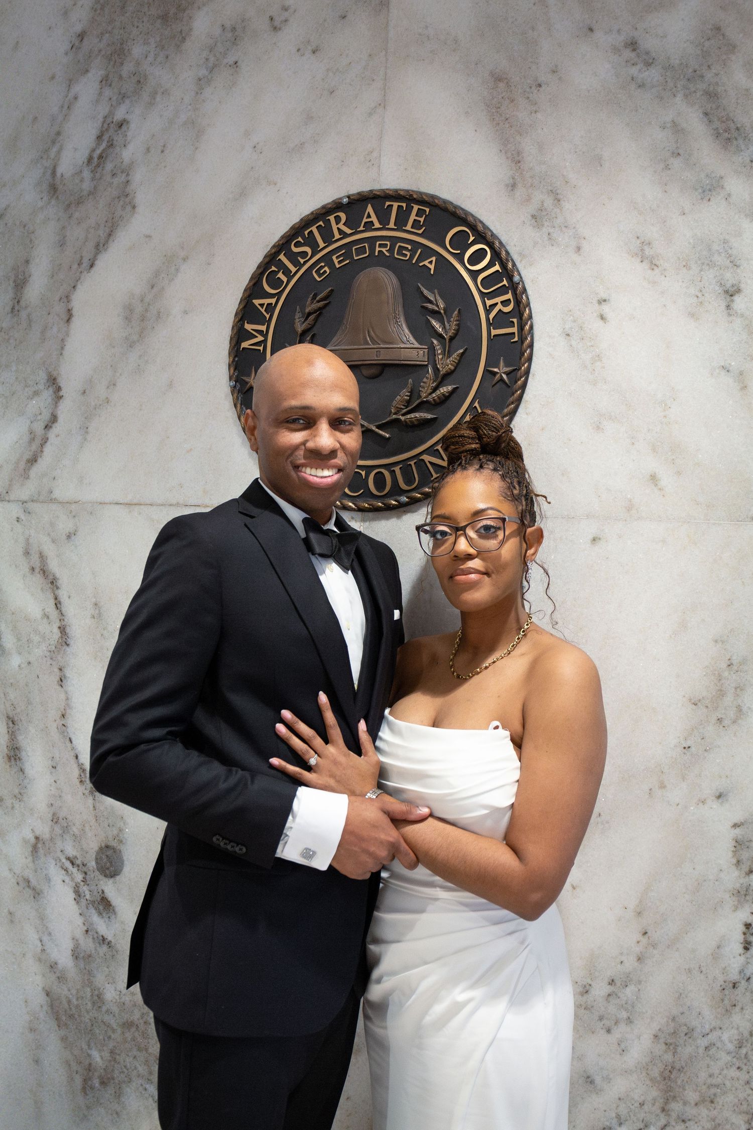 A couple poses together against a marble wall backdrop in formal wedding attire.