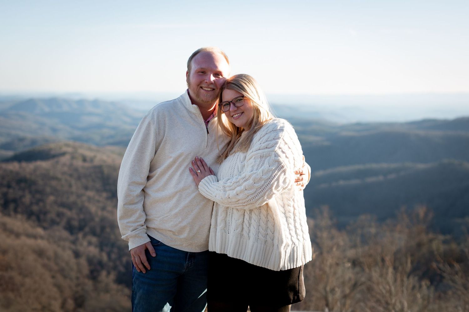 A couple embraces while standing on a scenic mountain overlook with rolling hills and forest in the background.
