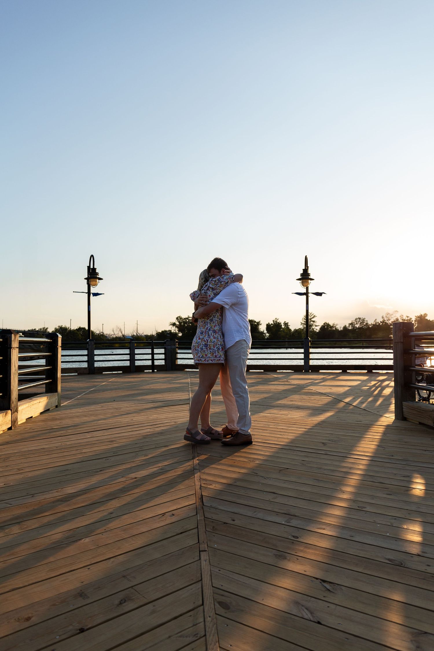 A couple embraces on a wooden pier at sunset with decorative lampposts and water in the background.