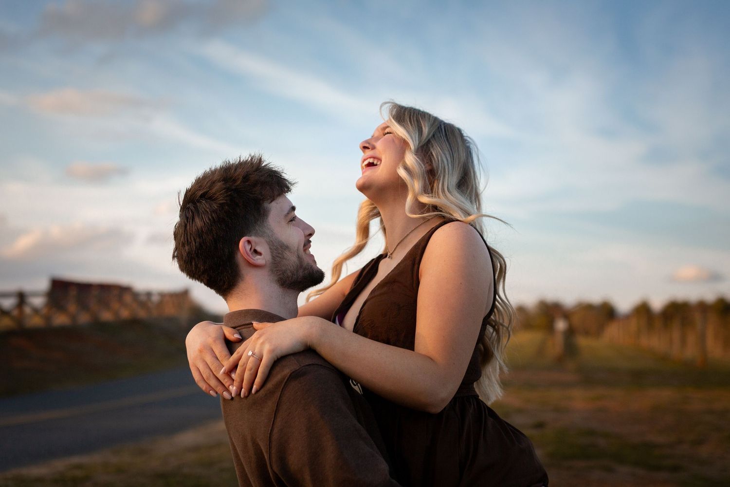 A joyful couple embraces outdoors at sunset, the woman laughing as the man lifts her against a blue sky backdrop.