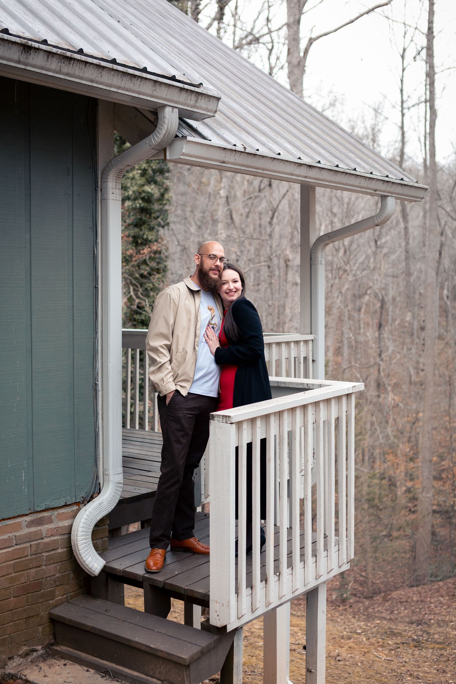 A couple stands together on a rustic covered porch with white railings during autumn in the woods.
