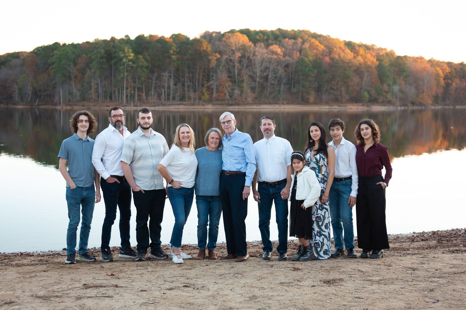 A large group stands together by a serene lake with autumn foliage reflecting in the calm water.