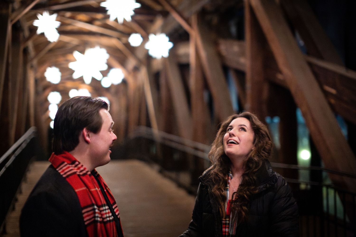 Two people share a joyful moment together while walking through a wooden covered bridge structure at night.
