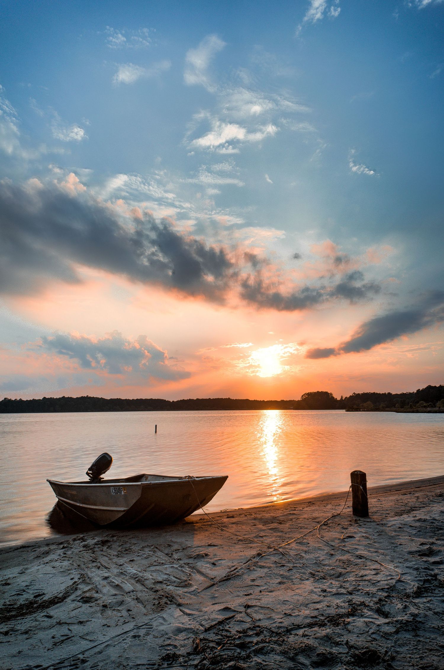 Aluminum motorboat resting on sandy beach at golden sunset with dramatic cloudy sky reflected on calm lake.