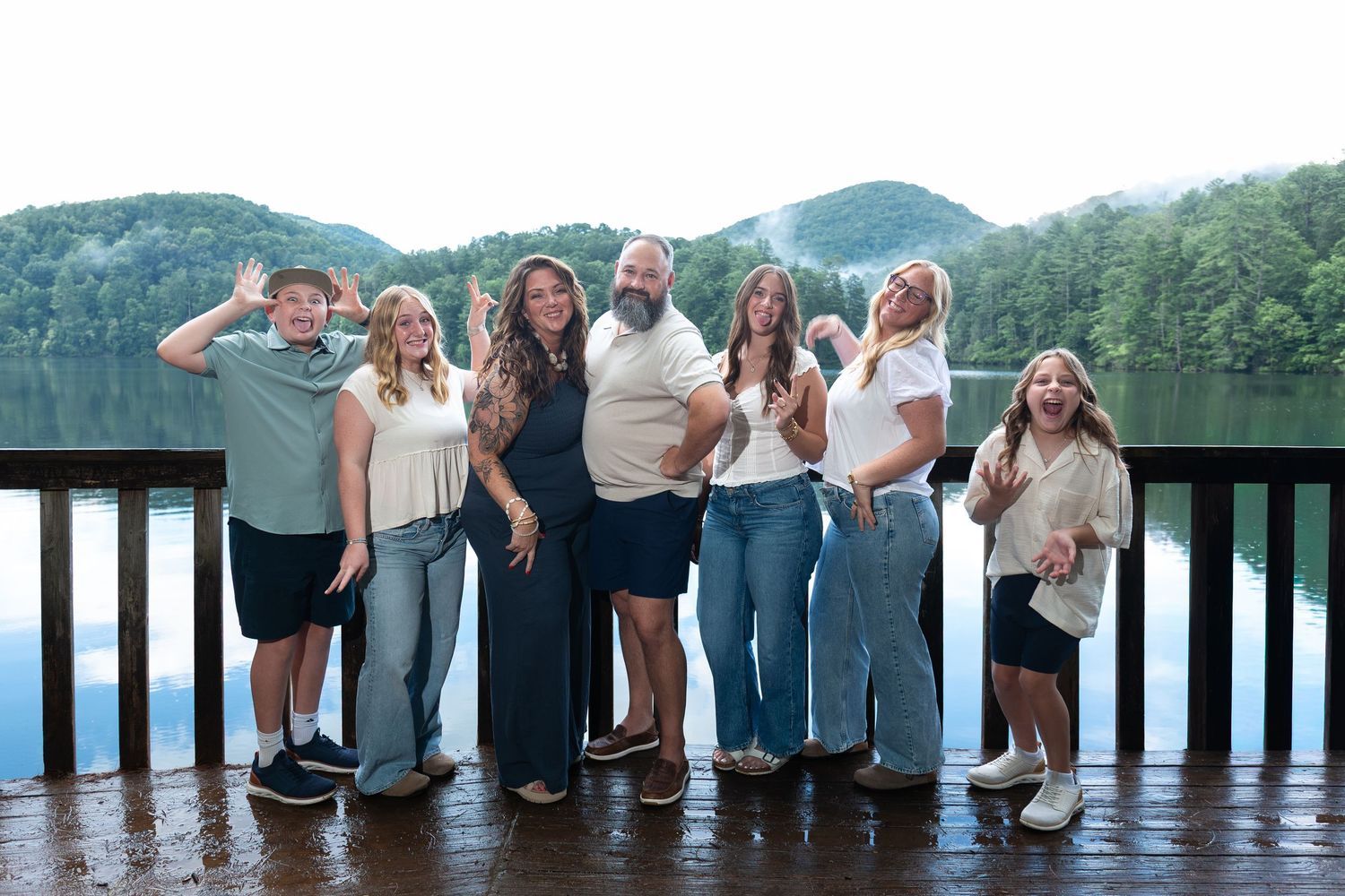 A cheerful group of seven people poses playfully on a wooden deck overlooking a misty lake and forested mountains.