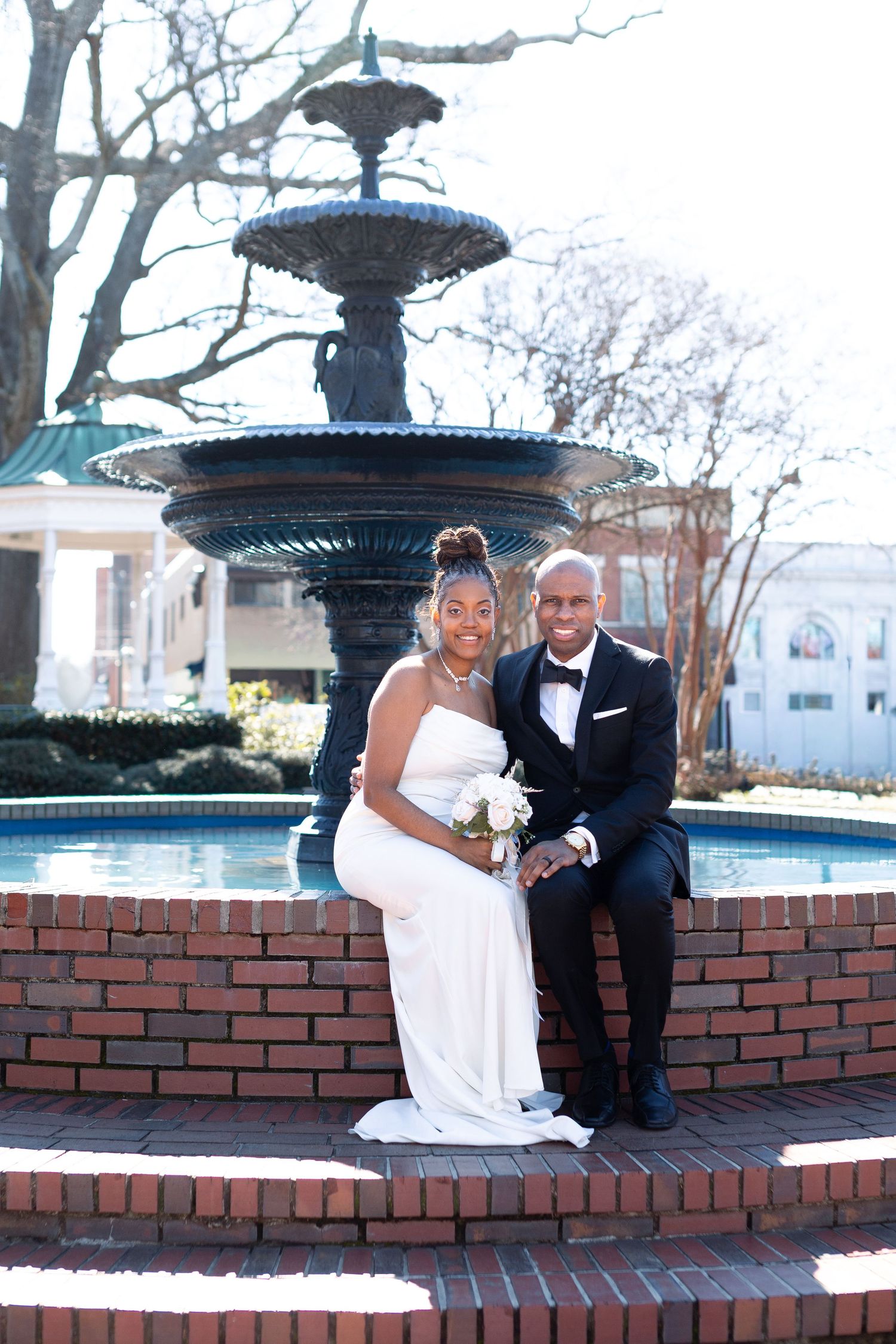 A newlywed couple sits on a brick ledge by an ornate fountain in a courtyard setting.