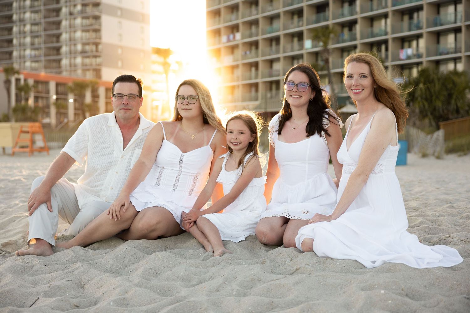 A group in white outfits sits together on a sandy beach at sunset with high-rise buildings in the background.