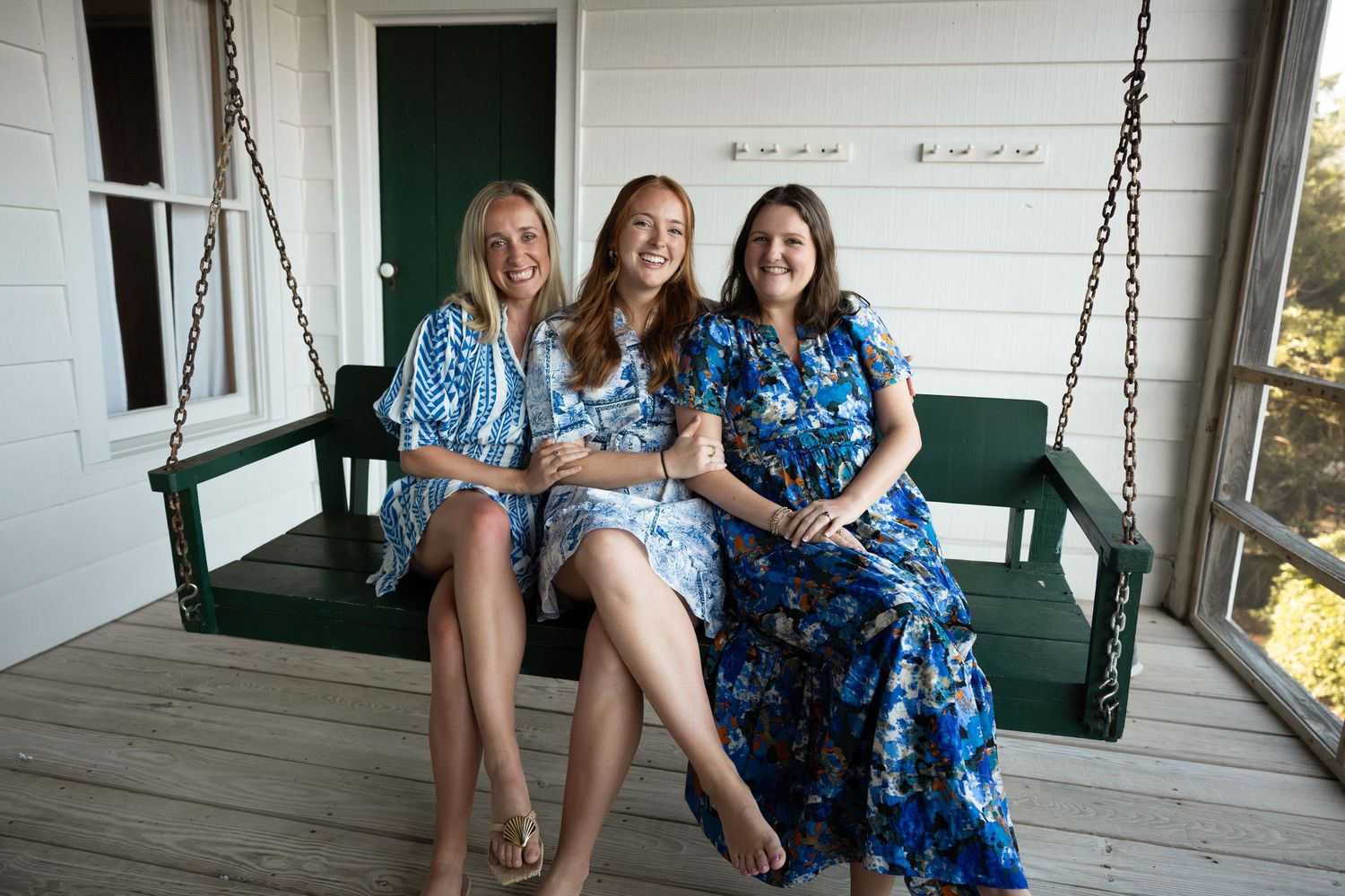 Three people in matching blue floral dresses sit together on a porch swing.