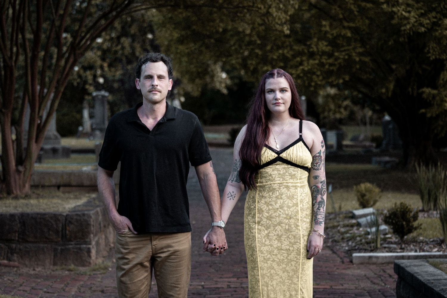 Couple holding hands in a cemetery, woman in yellow dress with tattoos, man in black shirt and khaki pants.