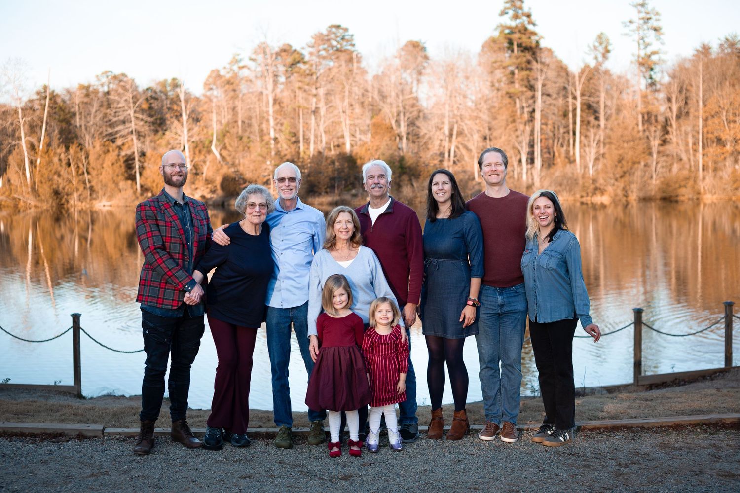 Multi-generational family group posing outdoors near a reflective lake surrounded by autumn trees at golden hour.
