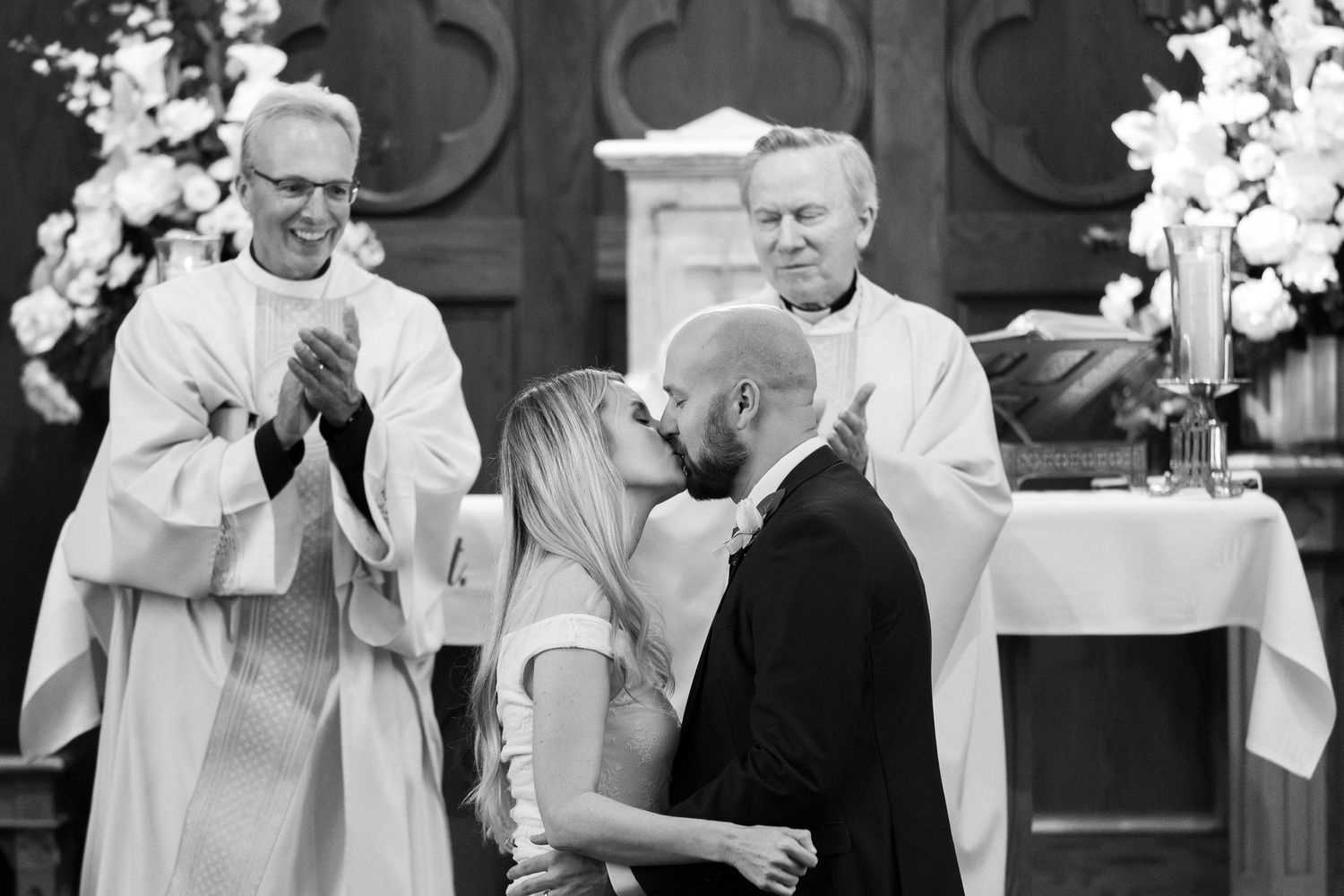Couple shares first kiss at church wedding altar while officiants applaud in black and white photo.