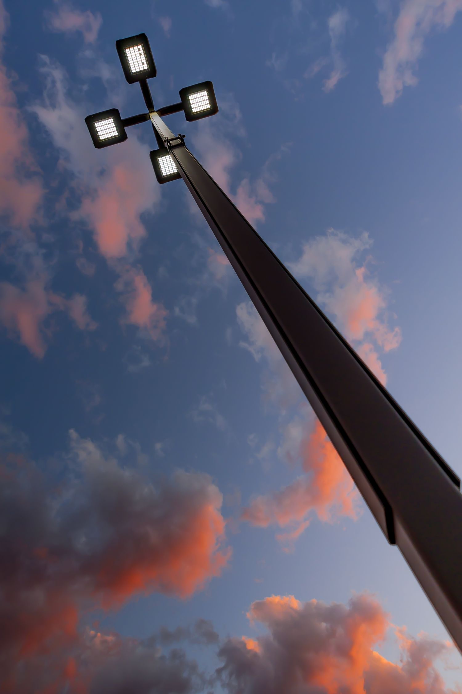 Low-angle view of a modern LED street light with four fixtures against a dramatic sunset sky with pink clouds.