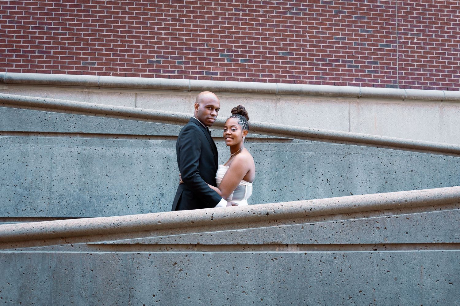 A romantic couple embraces along a curved railing with a brick building in the background.
