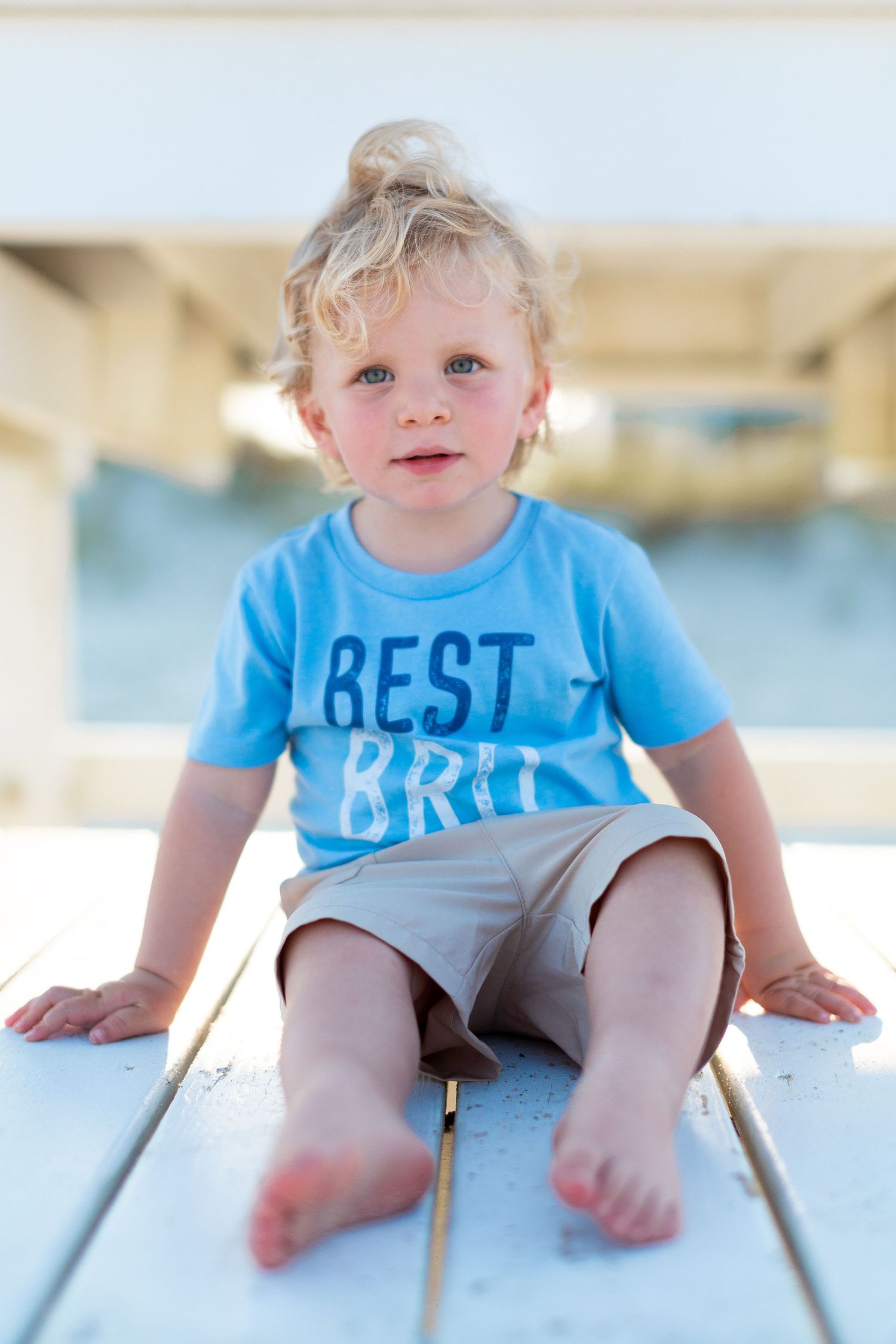 A toddler in a light blue 'Best Bro' t-shirt sits on wooden decking with bare feet.