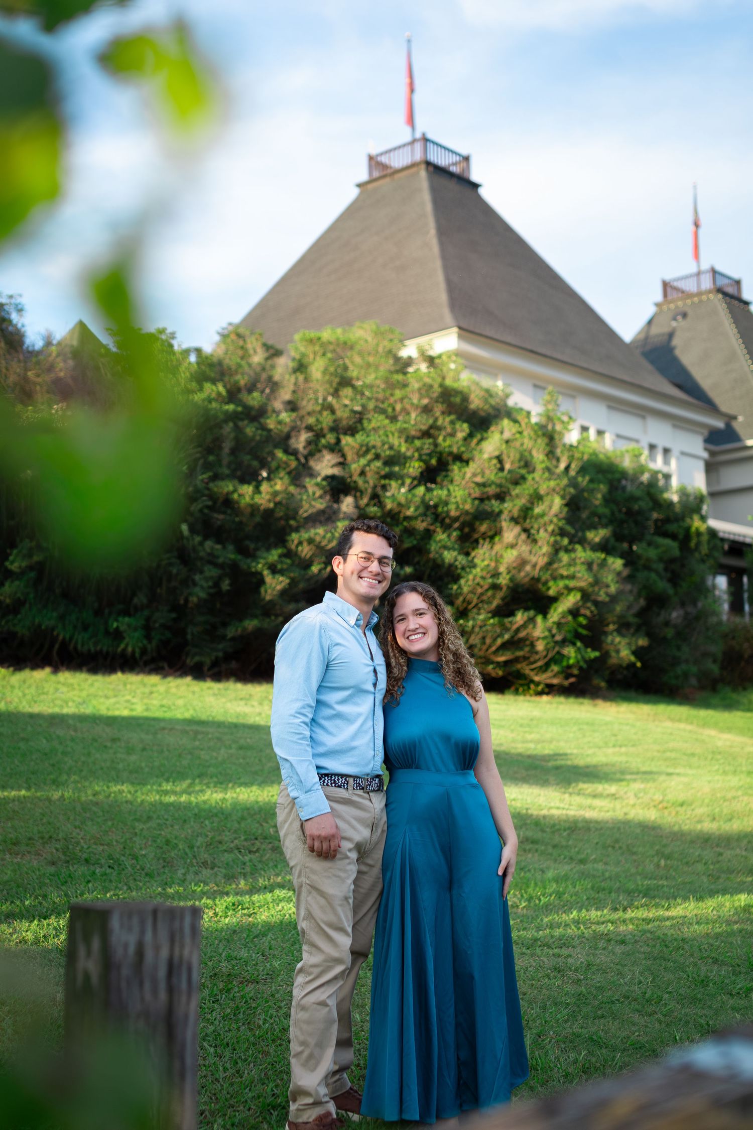 A smiling couple poses on green grass in front of a pyramid-roofed building surrounded by lush trees on a sunny day.