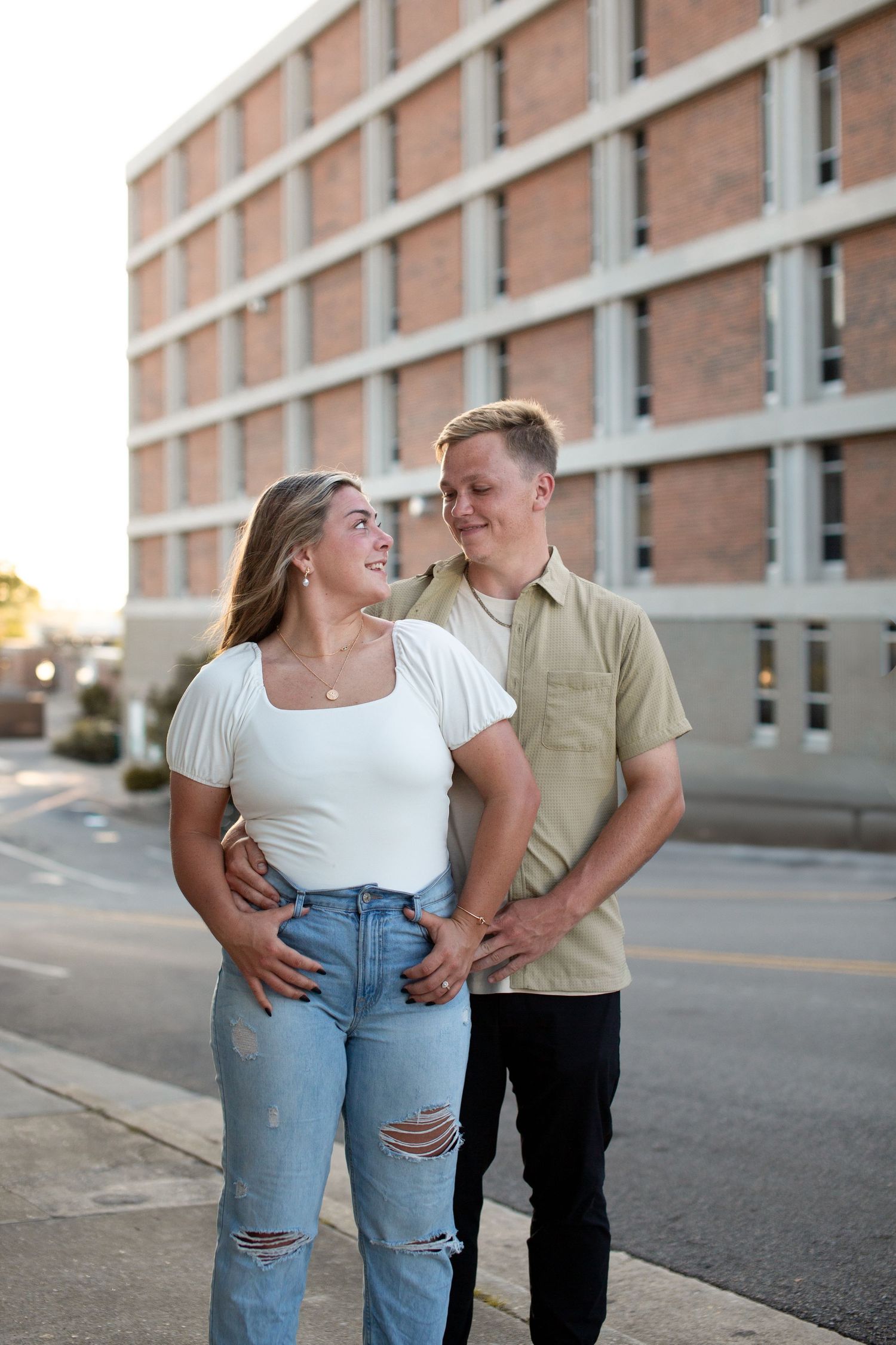 A young couple smiles at each other on a city sidewalk, woman in white top and ripped jeans, man in sage shirt.