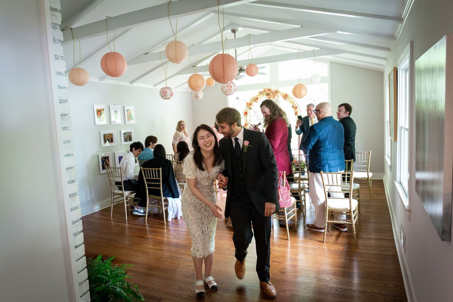 Smiling bride and groom walk hand in hand down a sunlit hall as wedding guests mingle beneath hanging coral paper lanterns.