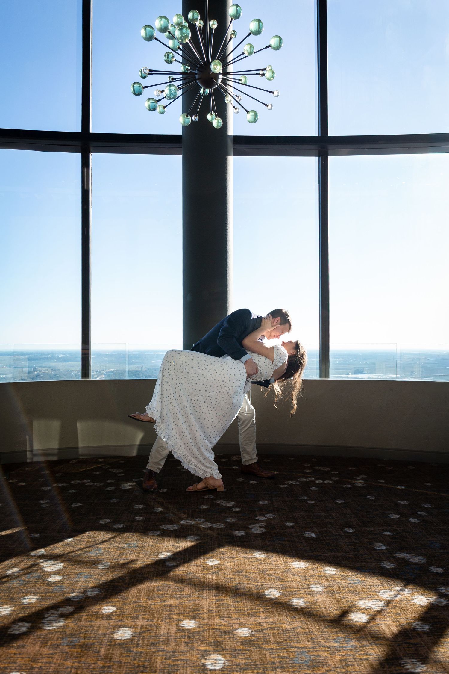 A romantic silhouette dances in front of large windows under a modern chandelier, creating dramatic lighting patterns on the floor.