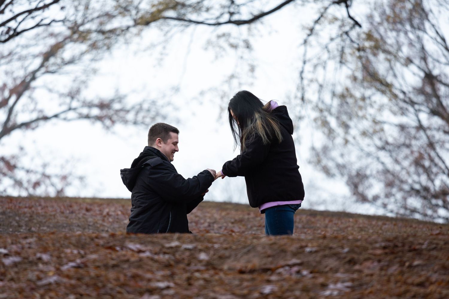 Two people stand on a hill against a cloudy sky while trees sway in the background.