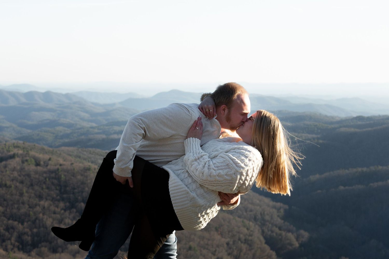 Two people share a romantic moment on a mountaintop overlooking a scenic valley landscape at sunset.