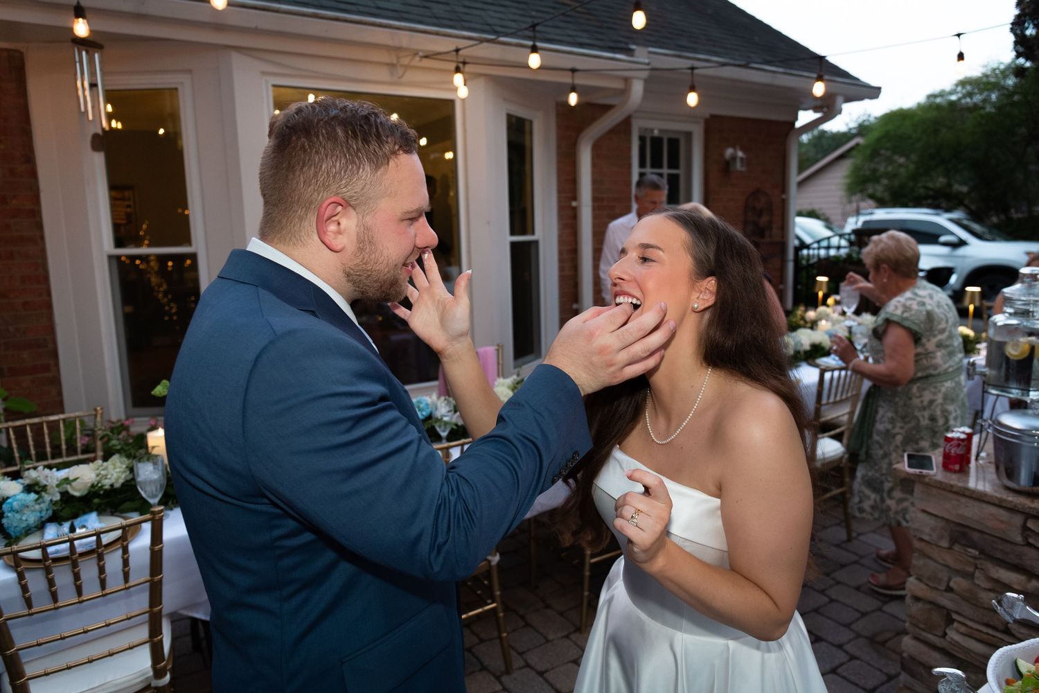 A groom in a navy suit playfully feeds wedding cake to his bride in a white dress during an outdoor reception with guests nearby.