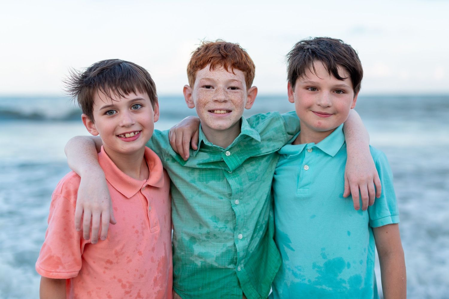 Three smiling boys in colorful polo shirts stand arm in arm on the beach with ocean waves behind them.