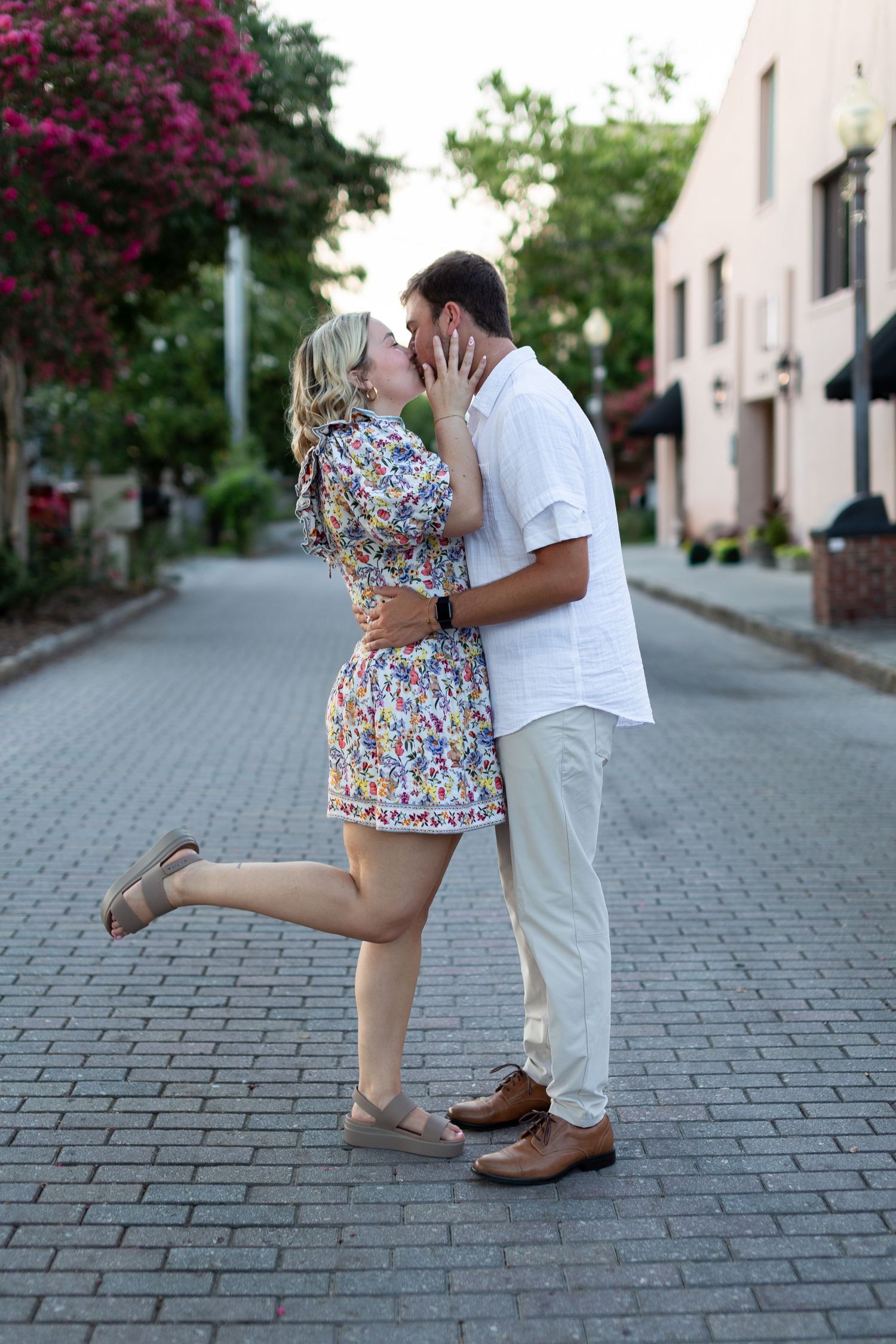 A couple embraces romantically on a brick paved street with pink flowers and buildings in the background.