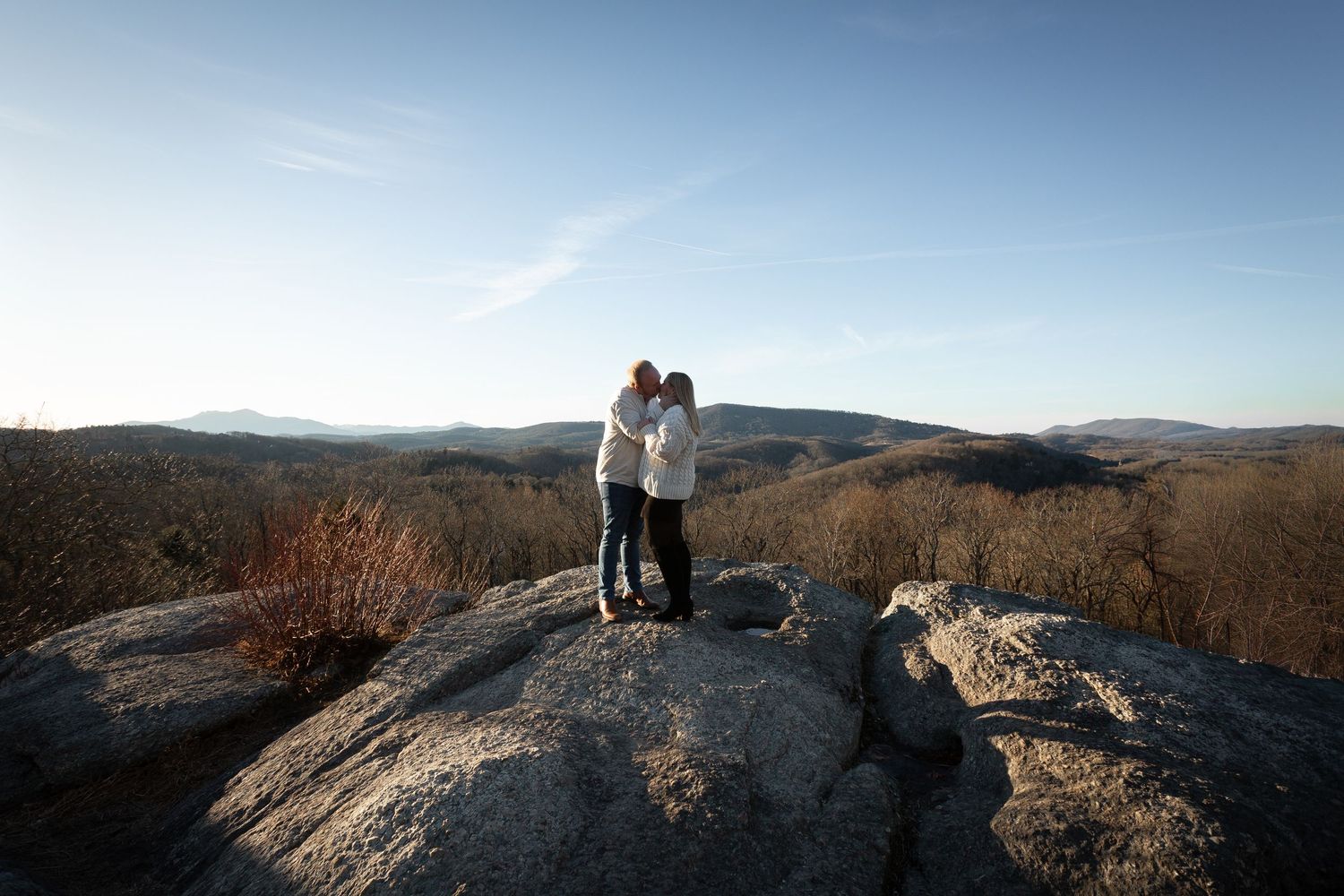A couple embraces while standing on a rocky overlook with rolling mountains and blue skies in the background.