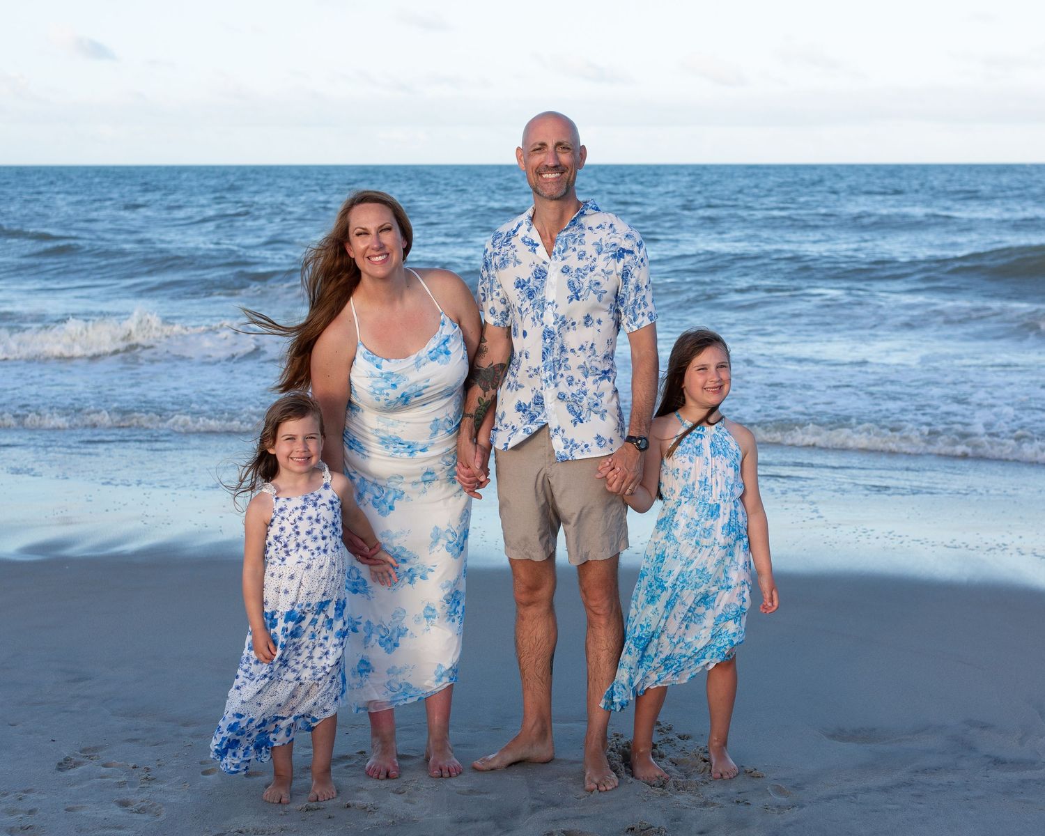 Family group wearing coordinated blue and white outfits on beach at sunset.