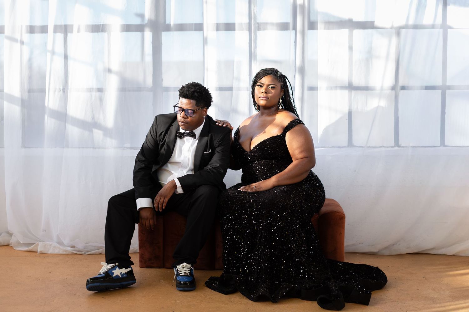 Two people in formal attire sit together on brown bench against white curtain backdrop at elegant event.