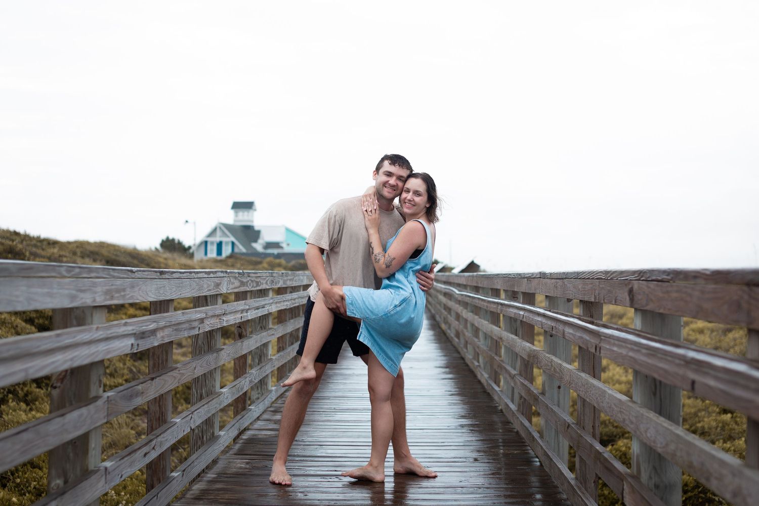 A couple embraces playfully on a wooden boardwalk with a blue domed building visible in the background.