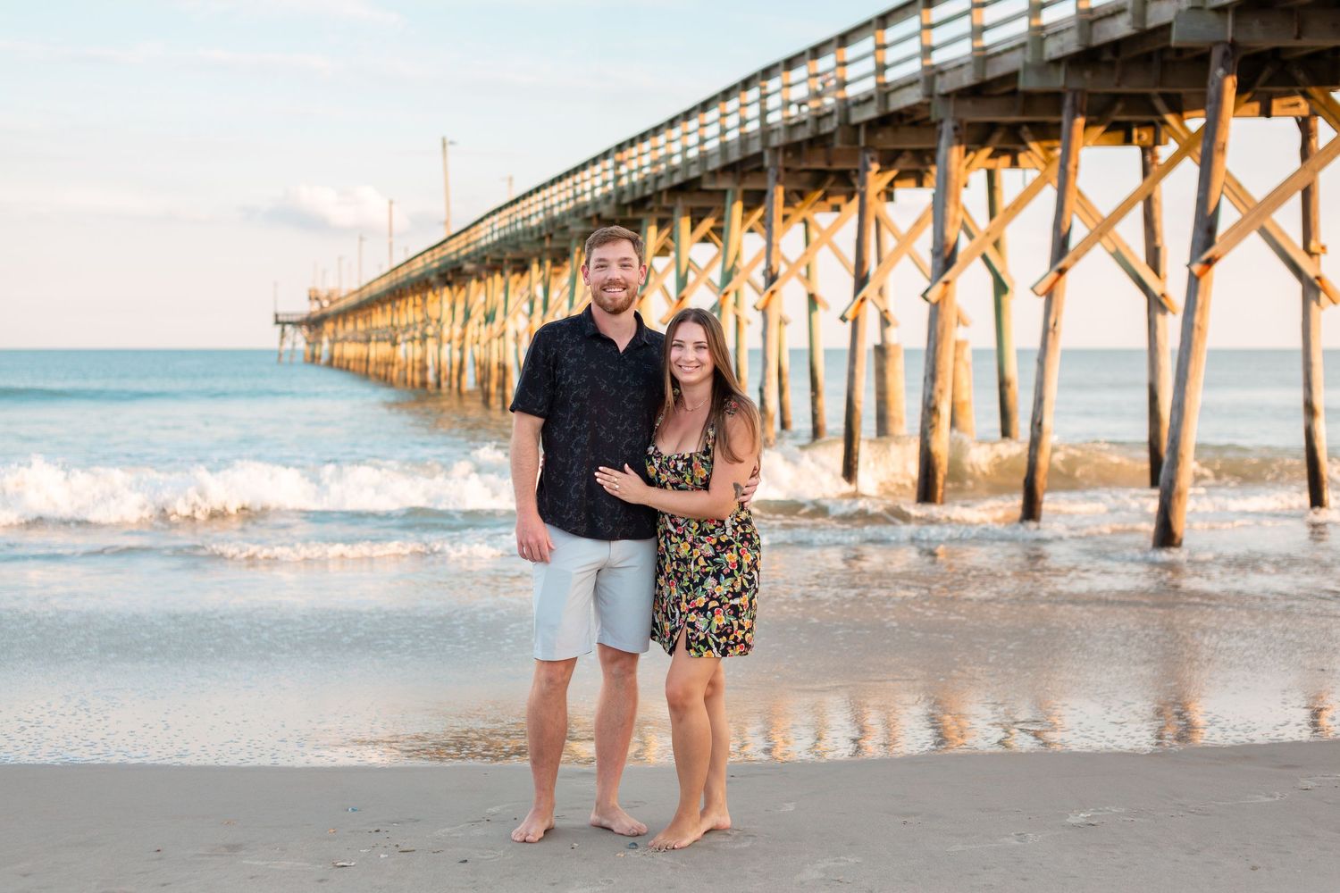 A couple poses together on the beach with a wooden pier in the background.