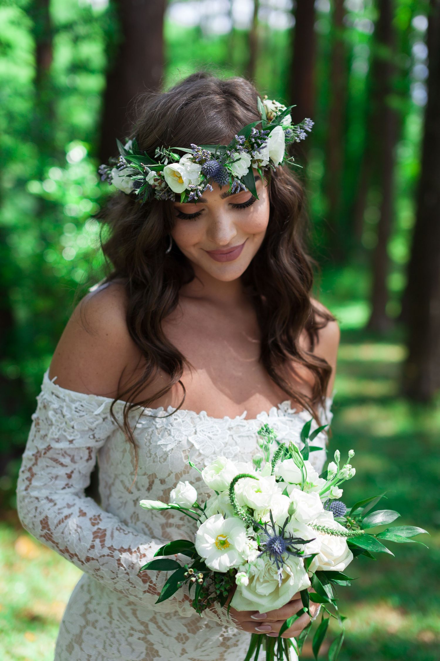 Bride in lace dress holds white floral bouquet while posing in a sunlit forest setting.